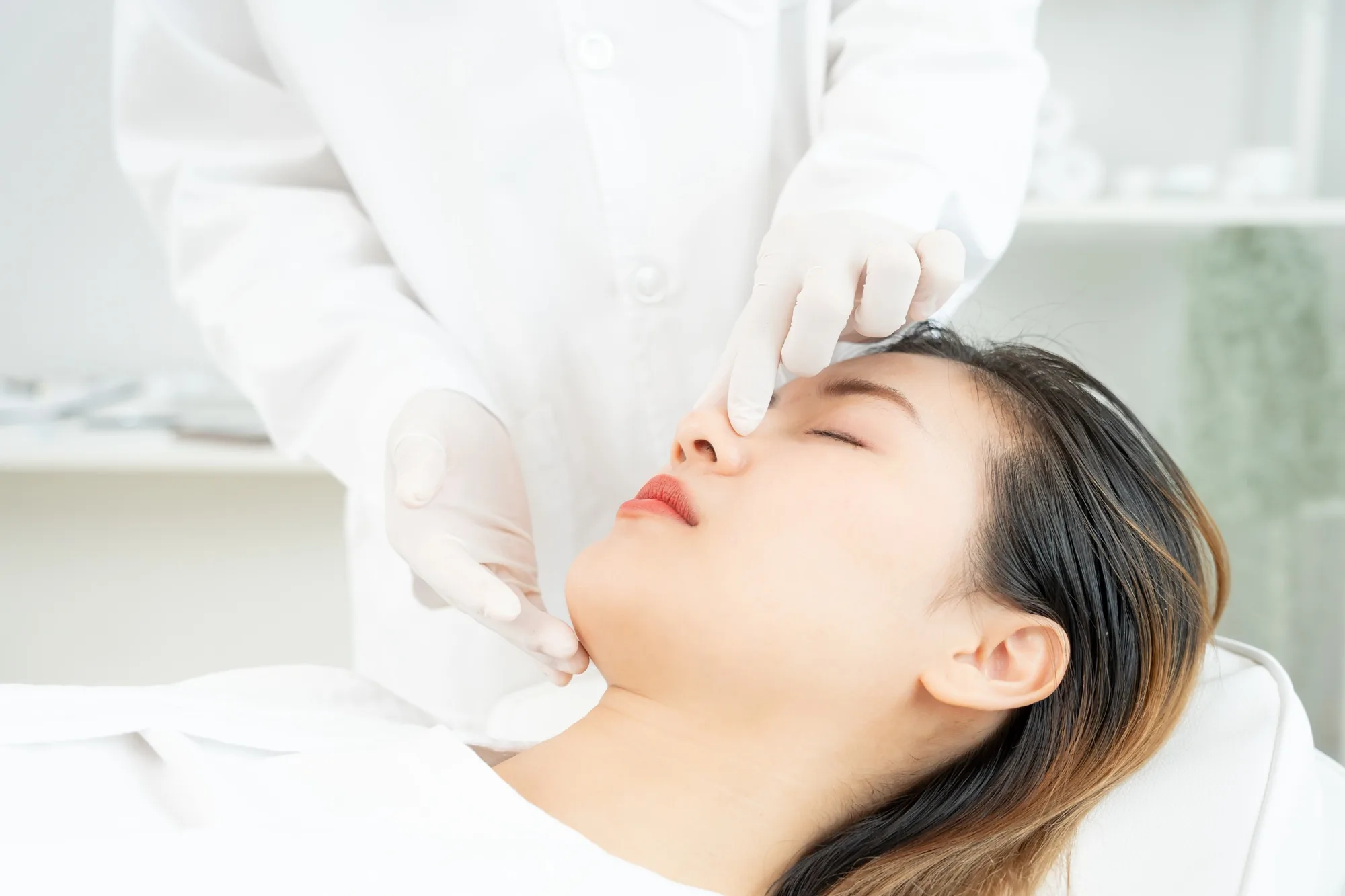 Doctor in white gloves examining a woman's nose and chin before a procedure